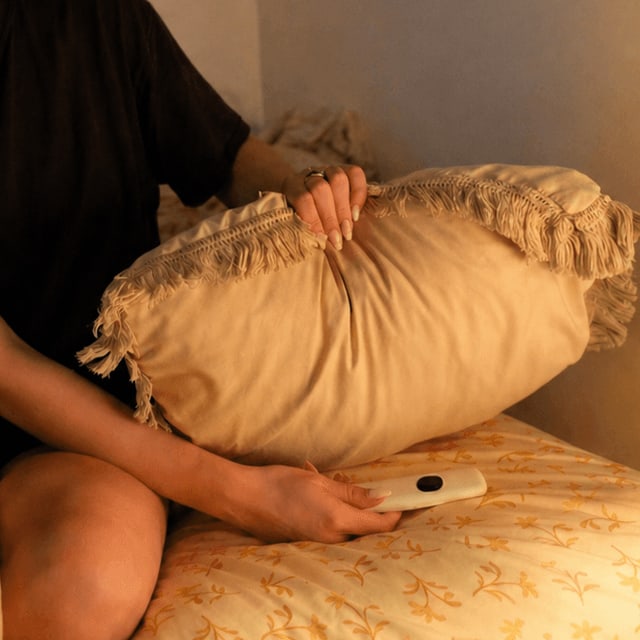 One hand lifting the edge of a pillow, the other holding the Lullabar beside it, ready to slide it under.