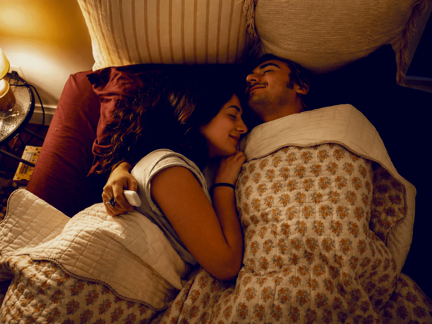 A couple asleep together in a warmly lit bedroom at night, one holding a slim pillow speaker